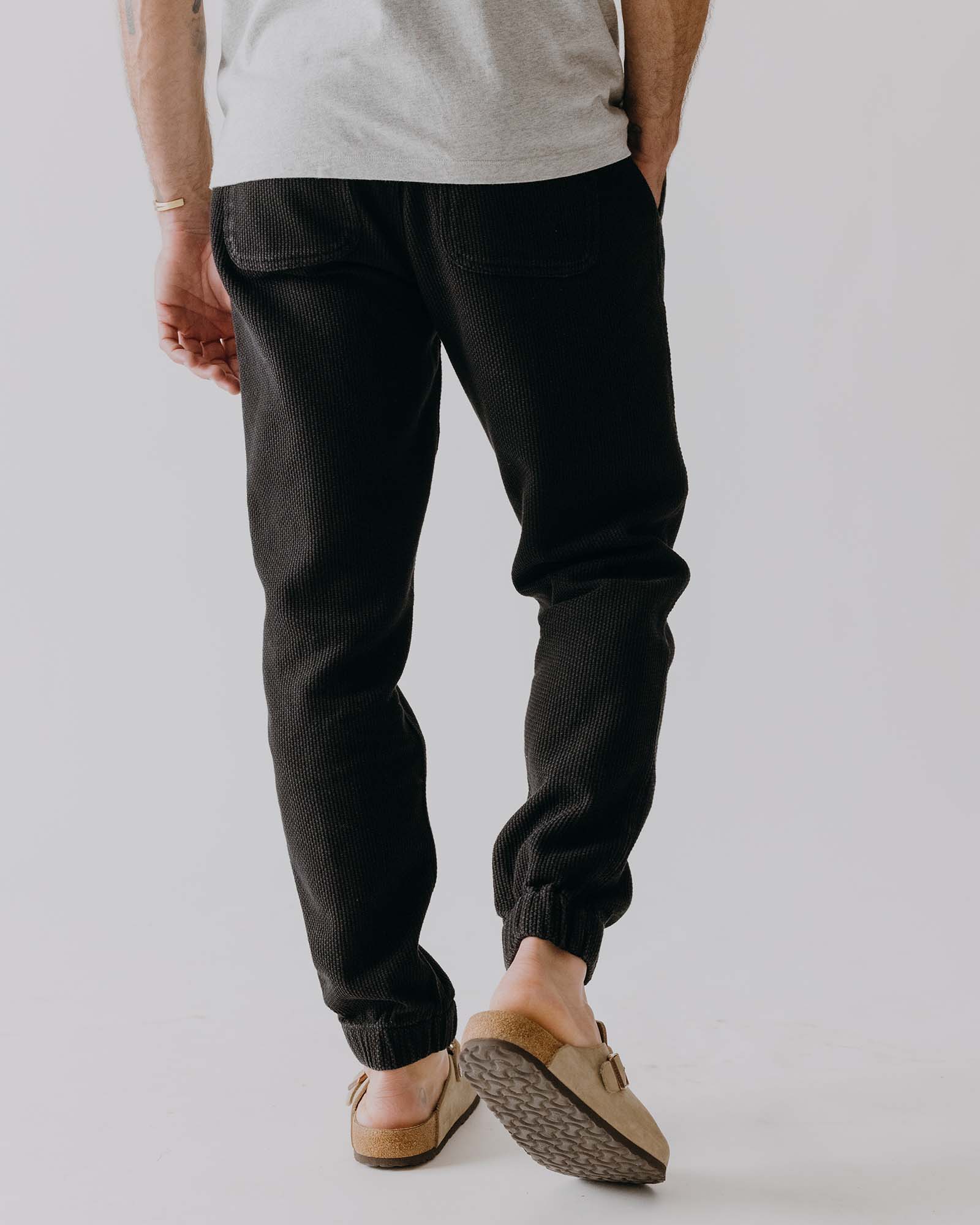 Person wearing black pants and beige shoes on a plain background, shown from the back