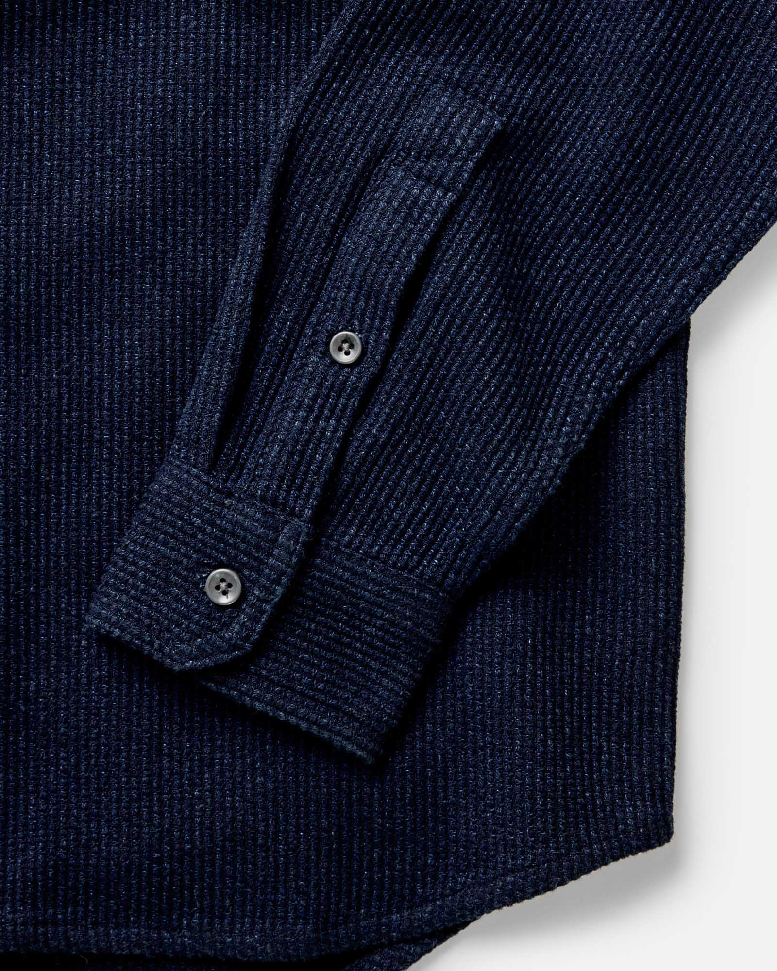 Close-up of Taylor Stitch Utility Shirt in Rinsed Indigo Sashiko showing buttoned cuff and sleeve detail with dark corozo-style buttons and pronounced horizontal sashiko weave texture on deep rinsed indigo organic cotton
