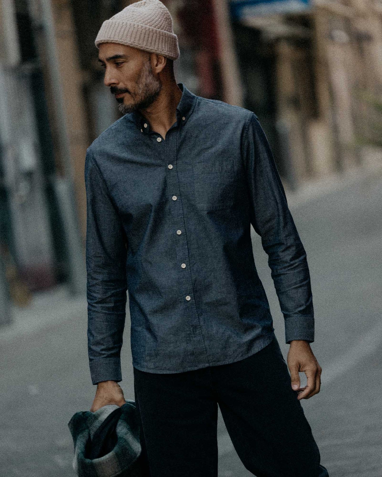 The Jack in Rinsed Indigo Chambray by Taylor Stitch, lifestyle front view, model wearing the shirt buttoned up with a beanie and dark trousers, walking outdoors on a cobblestone street