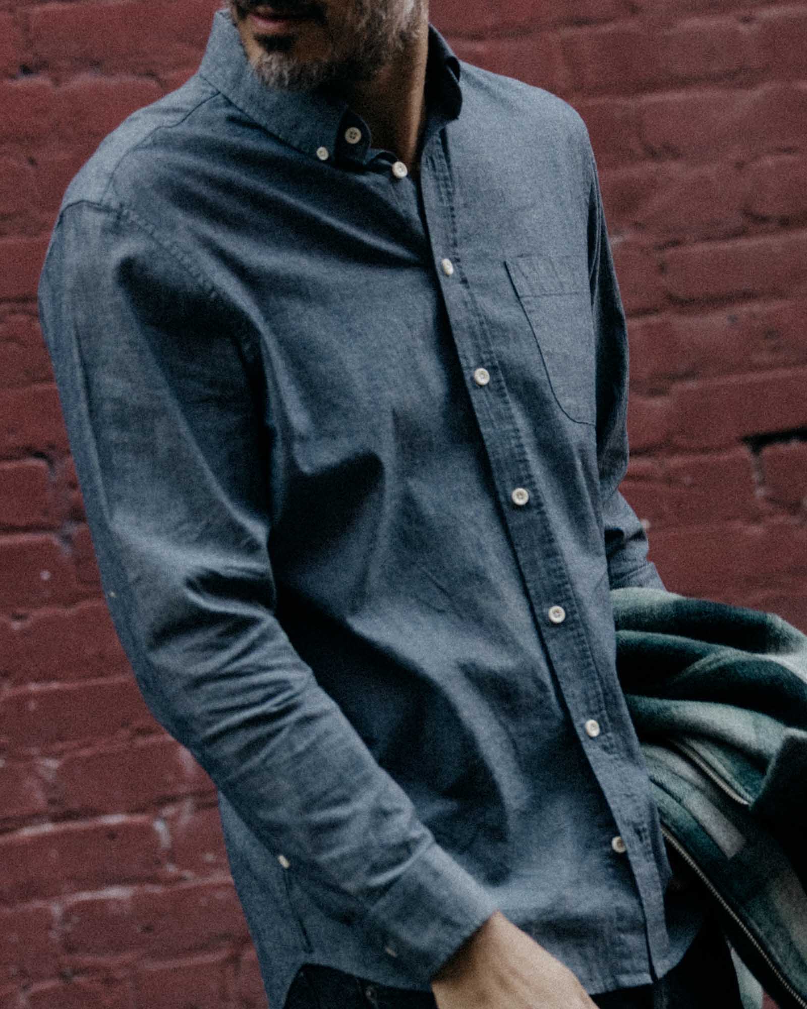 The Jack in Rinsed Indigo Chambray by Taylor Stitch, close-up front view showing button-down collar, chest pocket, and soft indigo chambray fabric texture against a red brick wall