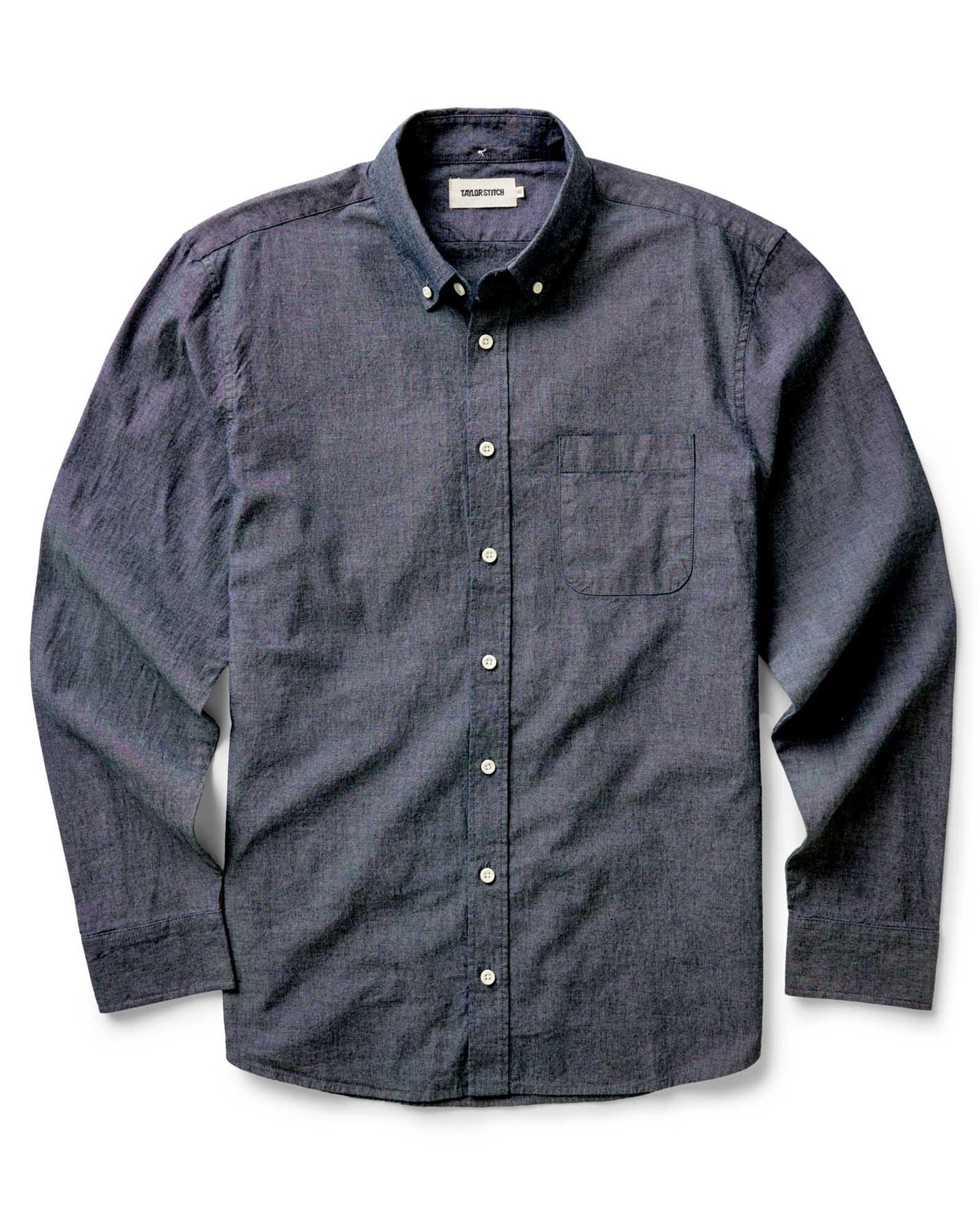 The Jack in Rinsed Indigo Chambray by Taylor Stitch, flat lay front view showing full shirt silhouette, button-down collar, chest pocket, and Taylor Stitch label on a white background