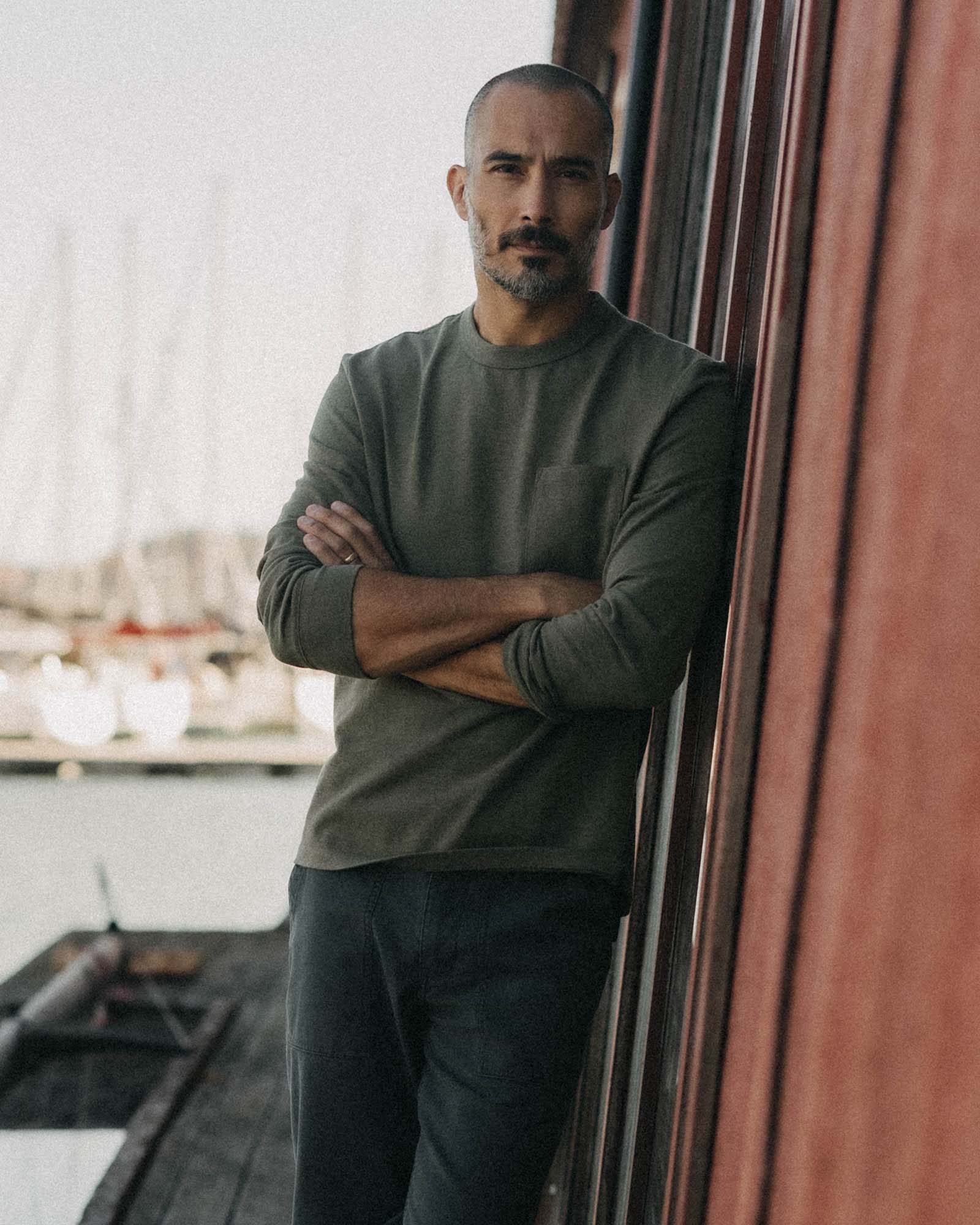 Man wearing The Long Sleeve Shop Tee in Army Twist by Taylor Stitch, arms crossed, leaning against a red wall with a marina in the background