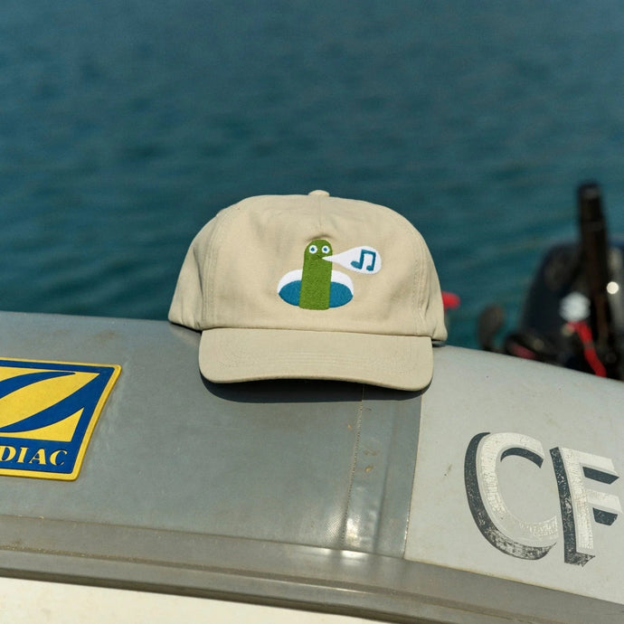 Beige cap with a green character on a boat with water in the background
