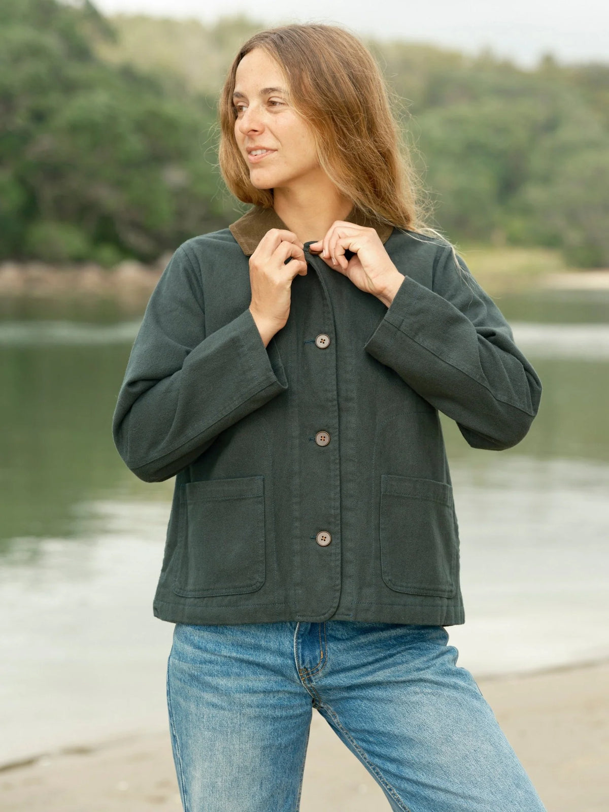 Front three-quarter view of a woman wearing the Mollusk Surf Shop Lined Painter Jacket in Black Forest, hands raised to the tan contrast collar, showing button front closure, two lower patch pockets, and a relaxed boxy silhouette in dark forest green 100% cotton, styled with light wash jeans at a lakeside
