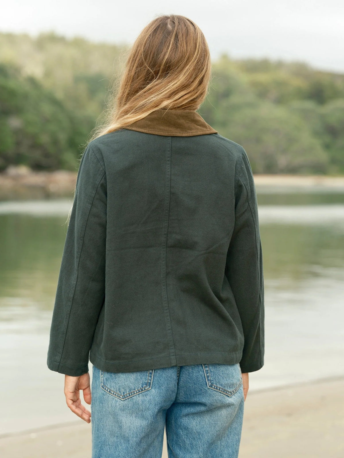 Back view of a woman wearing the Mollusk Lined Painter Jacket in Black Forest, showing the relaxed boxy cut, center back seam, long sleeves, and tan contrast collar detail in dark forest green 100% cotton, styled with light wash jeans at a lakeside with trees in the background
