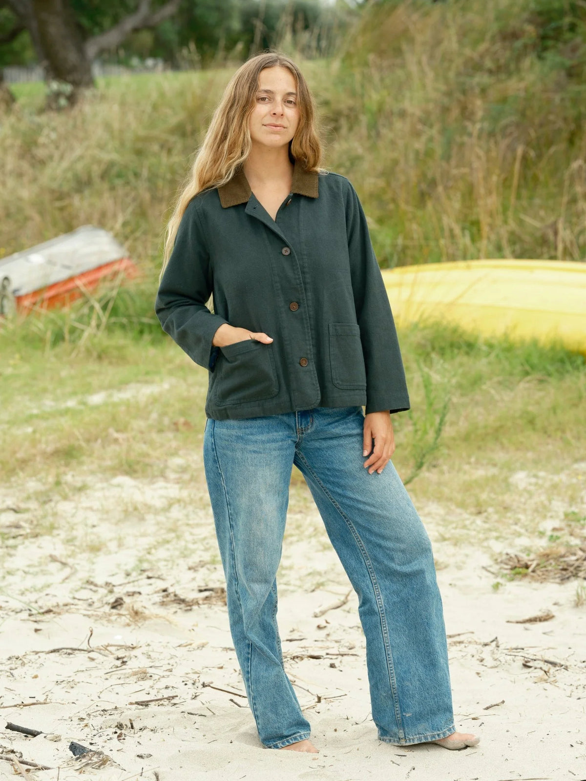 Full-length front view of a woman standing on a sandy beach wearing the Mollusk Lined Painter Jacket in Black Forest with wide-leg light wash jeans, hand in patch pocket, showing the relaxed boxy fit, button front, and tan collar against a grassy dune backdrop with colorful boats
