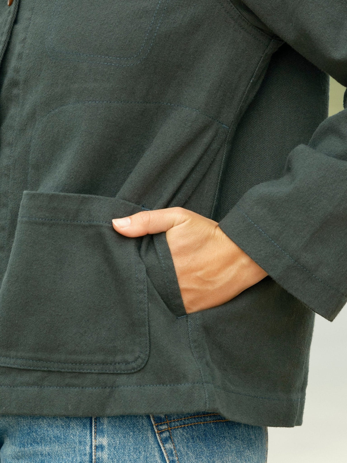 Close-up of the lower front of the Mollusk Lined Painter Jacket in Black Forest, showing a hand slipping into the hidden hand pocket above the lower patch pocket, highlighting the twill fabric texture, topstitched pocket edges, and long sleeve cuff in dark forest green 100% cotton
