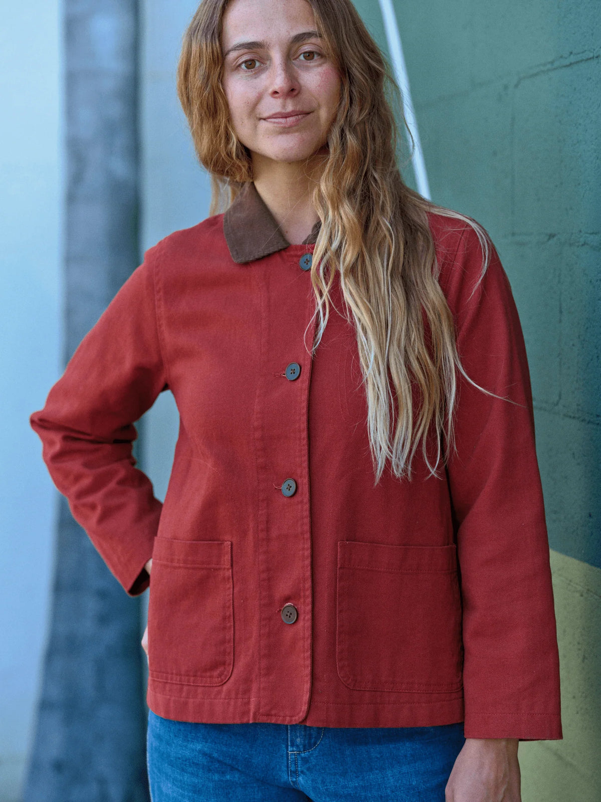 Front view of a woman wearing the Mollusk Surf Shop Lined Painter Jacket in Rust, showing button front closure with dark horn buttons, brown contrast collar folded down as a lapel, two lower patch pockets, and relaxed boxy fit in rust red 100% cotton, styled with blue jeans against a teal painted wall
