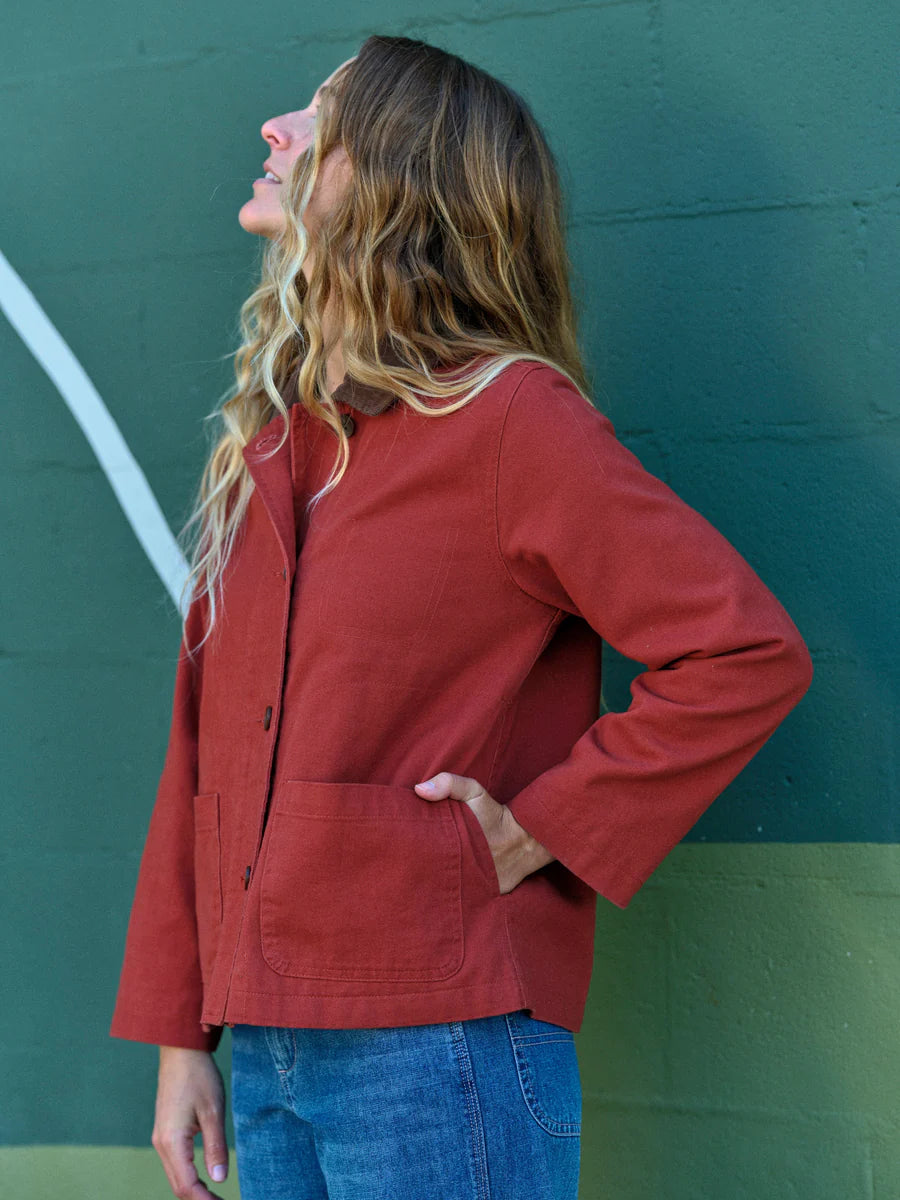 Side view of a woman wearing the Mollusk Lined Painter Jacket in Rust with hand in lower patch pocket, showing the relaxed boxy silhouette, long sleeves, side seam, and rust red 100% cotton fabric against a dark green painted wall
