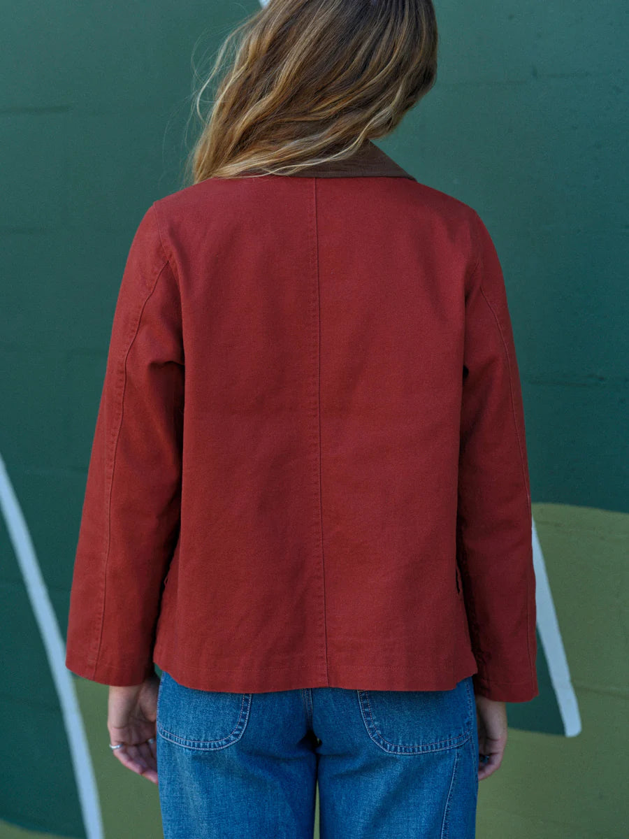 Back view of a woman wearing the Mollusk Lined Painter Jacket in Rust, showing the relaxed boxy cut, center back seam, and long sleeves in rust red 100% cotton against a dark green painted wall with white stripe detail
