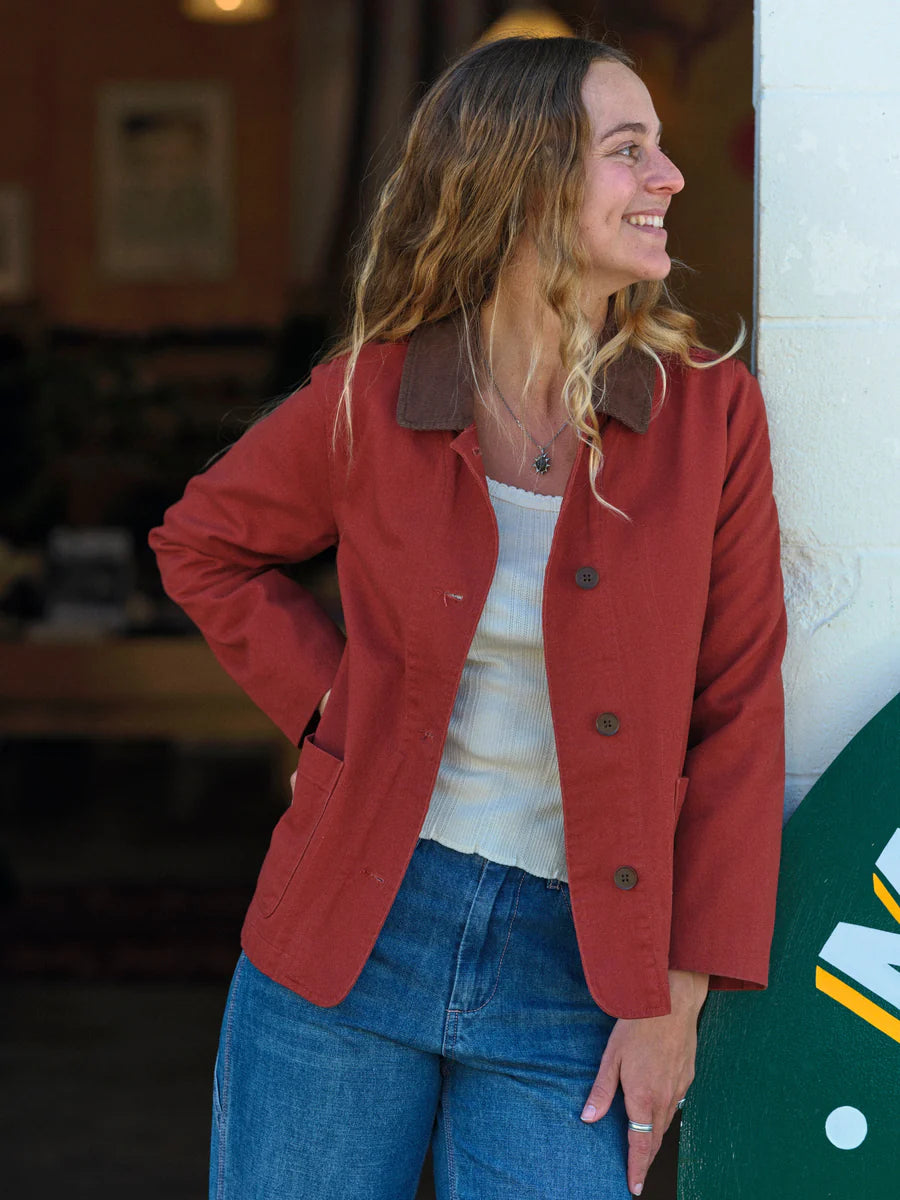 Three-quarter front view of a woman leaning against a wall wearing the Mollusk Lined Painter Jacket in Rust open over a white ribbed top, showing brown contrast collar, dark buttons, and relaxed boxy silhouette in rust red 100% cotton, styled with blue jeans in a warm indoor setting

