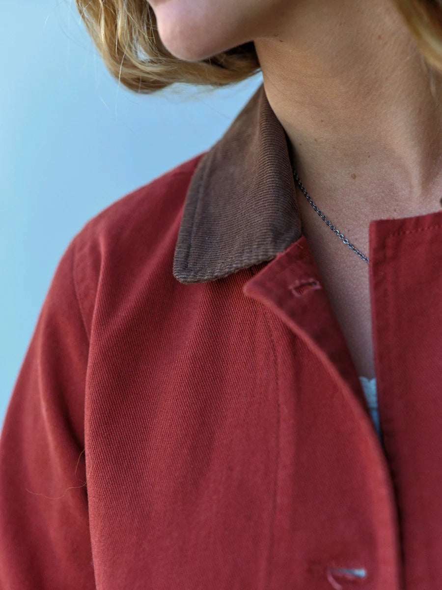 Close-up of the brown corduroy contrast collar of the Mollusk Lined Painter Jacket in Rust, folded open to show the lapel style, with rust red cotton twill body and a delicate silver chain necklace visible, against a light blue background
