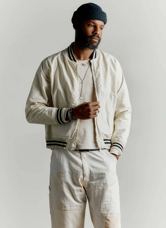 Man wearing The Madisonville Jacket by Imogene + Willie open over a cream mesh tee and white work pants, with a navy beanie and layered necklaces, showing the relaxed fit and black and white striped rib trim
