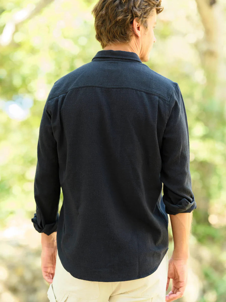 Model wearing Mollusk Surf Shop One Pocket Shirt in Black Flannel, back view walking in dappled outdoor light showing deep black brushed 95% cotton 5% linen flannel, point collar, clean back yoke, long sleeves rolled to mid-forearm, and slight saddle curve hem, styled with cream trousers
