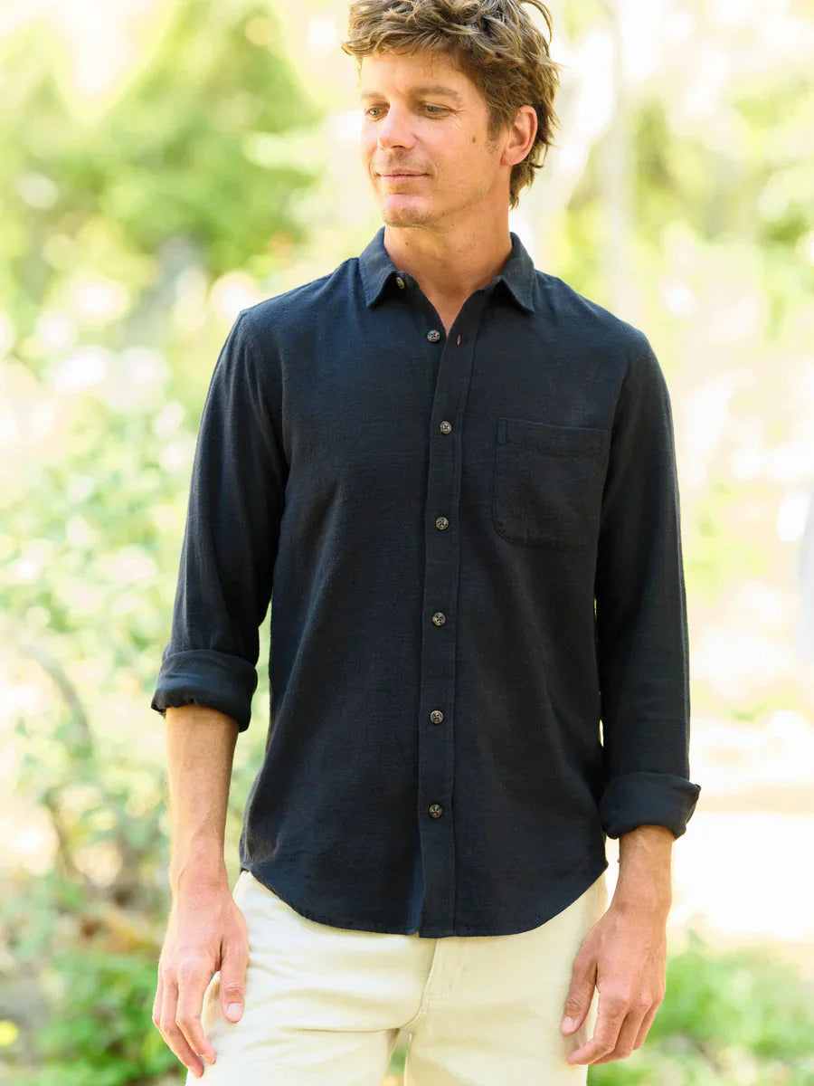 Model wearing Mollusk Surf Shop One Pocket Shirt in Black Flannel, full front view in dappled outdoor light with palm trees showing deep black brushed 95% cotton 5% linen flannel, point collar, long sleeves rolled to mid-forearm, tonal dark button-front placket, and single left-chest patch pocket, styled with cream trousers
