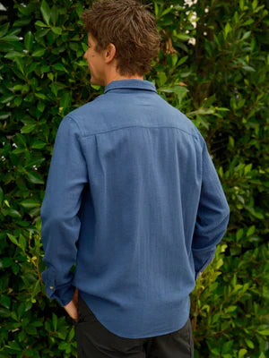 Model wearing Mollusk Surf Shop One Pocket Shirt in Indigo Flannel, back view against lush green hedge showing medium indigo blue cotton-linen flannel, point collar, long sleeves with button cuffs, clean back yoke, and slight saddle curve hem, styled with black trousers
