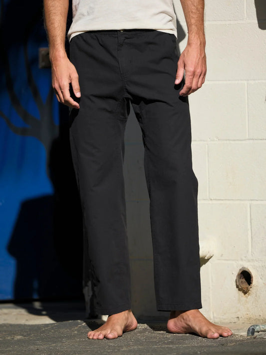 Model wearing the Mollusk Surf Shop Pinnacle Pants in Black, front view from waist down, showing relaxed straight leg, slight taper at ankle, and clean cropped hem of black cotton utility pant, styled with a white tee, barefoot on concrete
