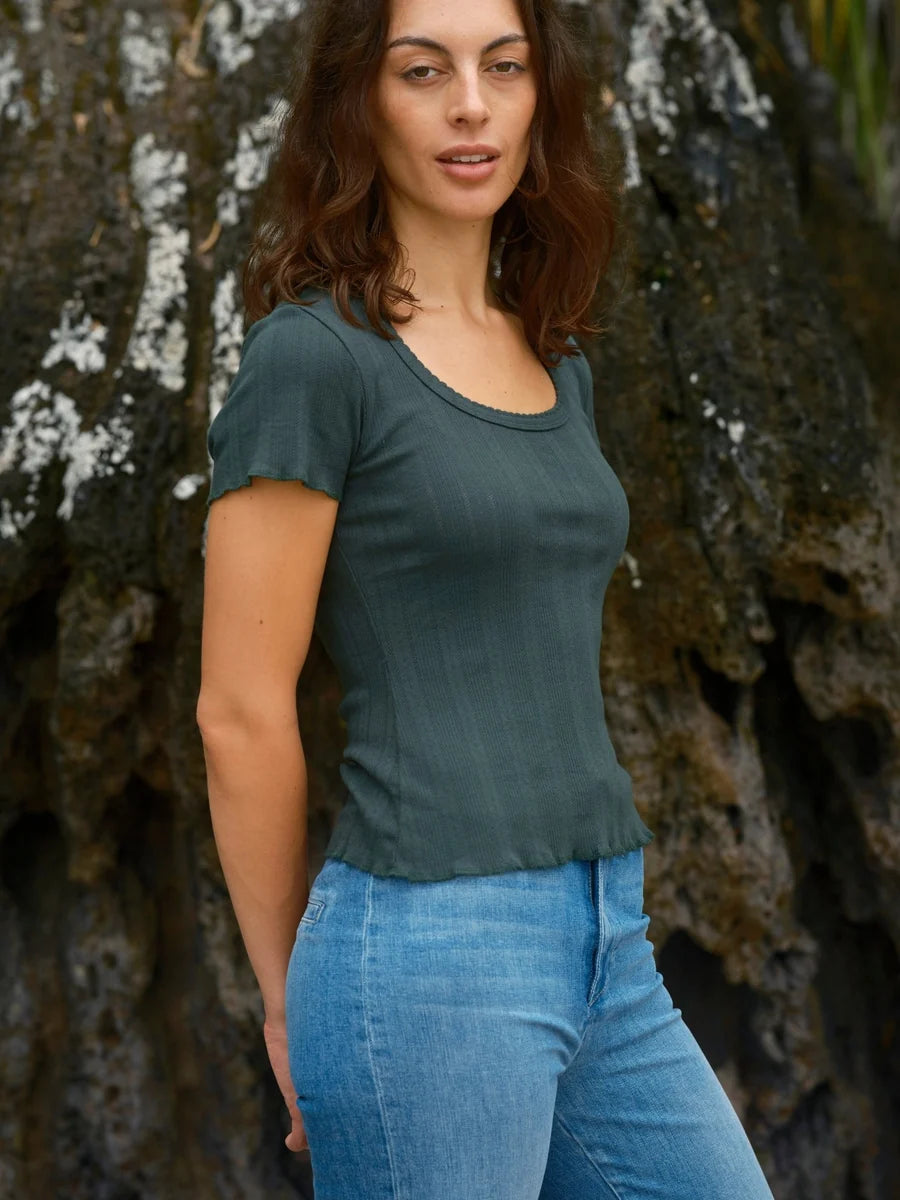 Female model wearing the Mollusk Surf Shop Pointelle Tee in Black Forest, three-quarter side view showing fitted silhouette, ruffled hem, and scoop neck, styled with light blue jeans outdoors