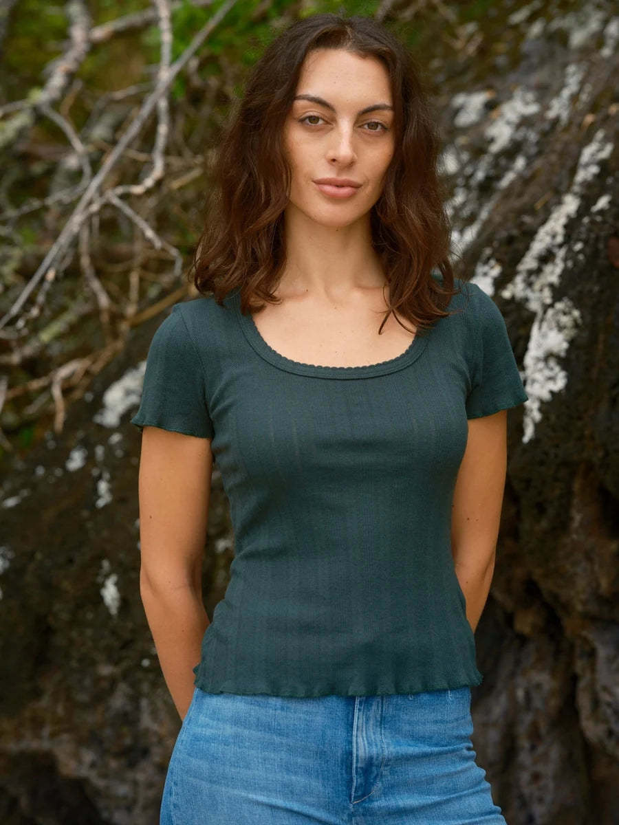 Female model wearing the Mollusk Surf Shop Pointelle Tee in Black Forest, a deep teal short-sleeve cotton tee with scoop neck and ruffled trim, styled with light blue jeans against a rocky outdoor backdrop