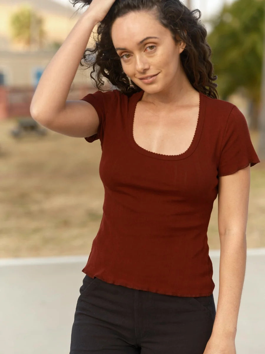 Female model wearing the Mollusk Surf Shop Pointelle Tee in Malbec, front-facing lifestyle shot showing scoop neck, ruffled trim, and fitted silhouette, styled with black pants outdoors