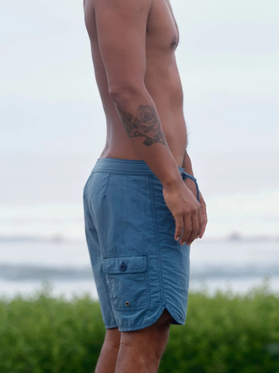 Male model wearing the Mollusk Surf Shop Scallop Trunks in Indigo, side profile showing indigo blue boardshorts with scallop curved hem, back cargo pocket with snap closure, photographed outdoors with ocean in background