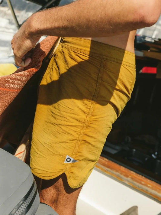 Mollusk Surf Shop Scallop Trunks in Mustard, close-up side view showing Velcro fly, back patch pocket, and pennant patch logo, worn on a boat in sunlight


