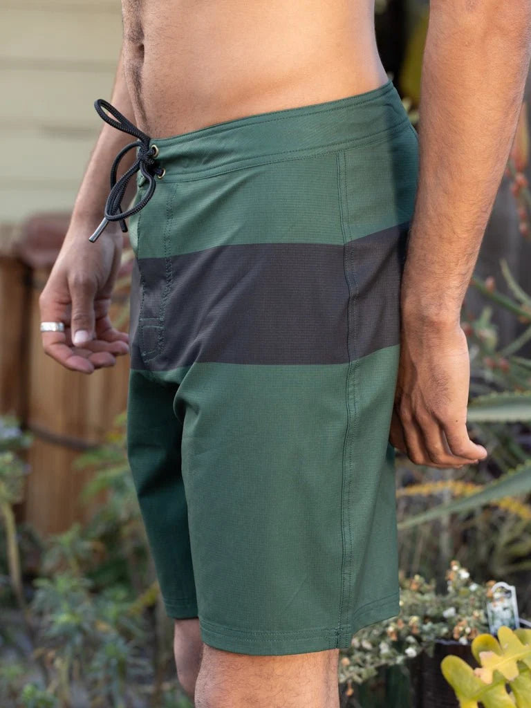 Male model wearing the Mollusk Surf Shop Stretch Notched Trunks in Black Rover Stripe, side view showing forest green and black stripe colorblock pattern, black lace-up drawstring, and notched hem, photographed outdoors
