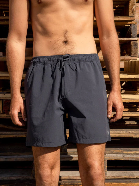 Male model wearing the Mollusk Surf Shop Stretch Vacation Trunks in Navy, front view showing navy nylon shorts with elastic waistband and drawstring, photographed outdoors against stacked wooden pallets in sunlight