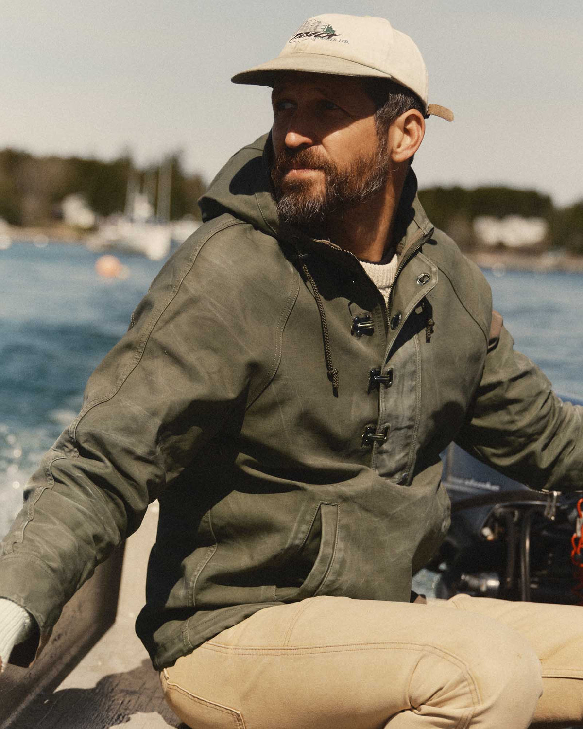 Model seated by the water wearing the fatigue olive Hatteras Anorak, showing the hood and kangaroo pocket.
