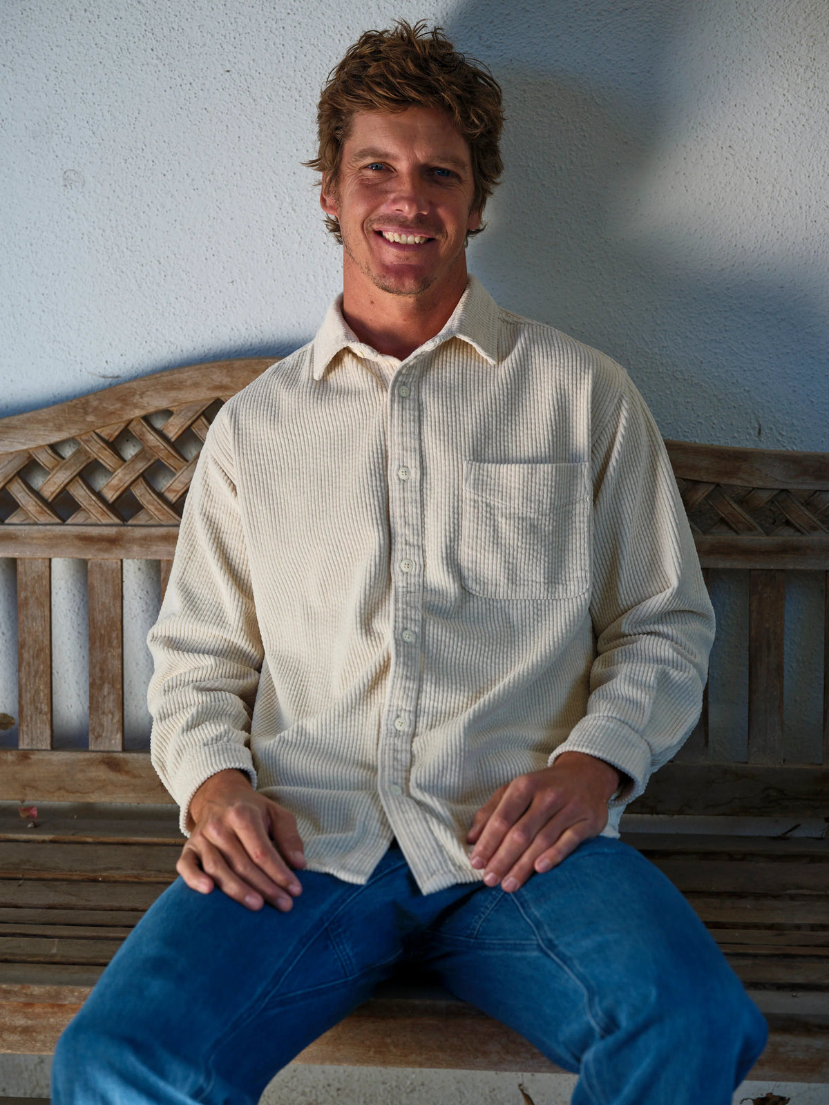 Front portrait of a smiling man seated on a weathered teak garden bench wearing the Mollusk Surf Shop Thurston Shirt in Natural Corduroy, showing the relaxed boxy fit, button front, point collar, and left chest patch pocket in cream-natural bubble corduroy, styled with medium blue jeans against a white stucco wall

