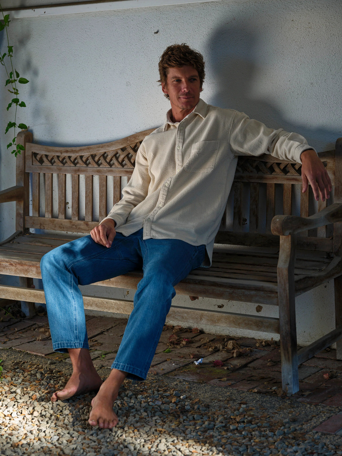 Full-length shot of a man seated barefoot on a weathered teak bench wearing the Mollusk Thurston Shirt in Natural Corduroy, showing the relaxed boxy silhouette, long sleeves, and cream-natural bubble corduroy fabric, styled with medium blue jeans on a gravel patio beside climbing foliage
