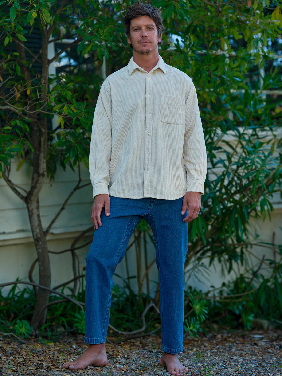 Full-length front view of a man standing barefoot in a garden wearing the Mollusk Thurston Shirt in Natural Corduroy, showing the relaxed boxy fit, button front, point collar, and chest pocket in cream-natural 100% cotton bubble corduroy, styled with medium blue jeans surrounded by lush green foliage
