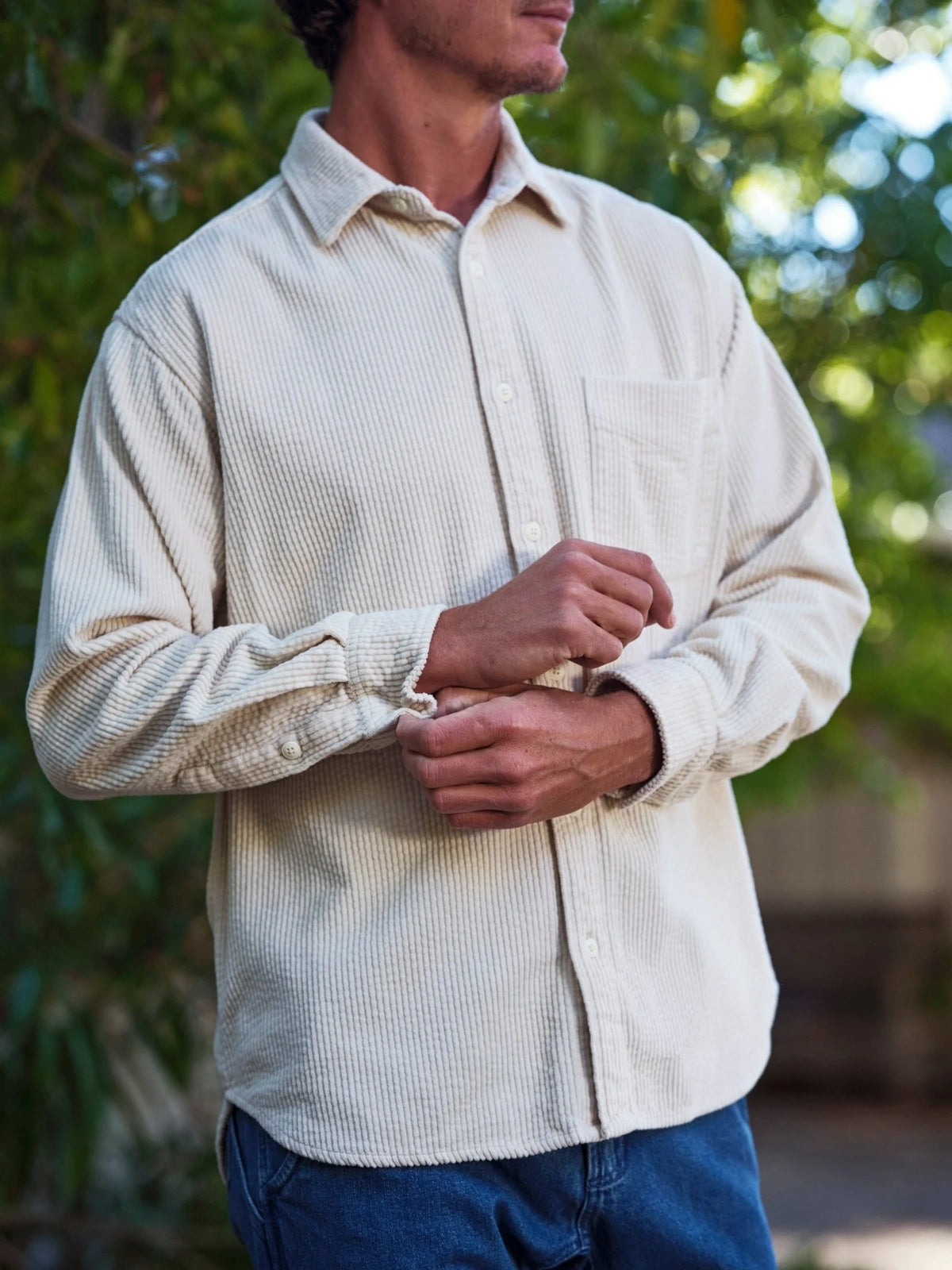 Close-up of a man rolling up the cuff of the Mollusk Thurston Shirt in Natural Corduroy, showing the wide bubble corduroy wale texture of the twisted-yarn weave, button placket, and cuff buttons in cream-natural 100% cotton, with a green garden in soft focus behind
