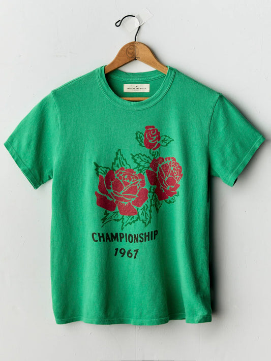 A green short-sleeve t-shirt hanging on a wooden hanger against a light gray wall. The shirt features a graphic print of three red roses with green leaves and the text 'CHAMPIONSHIP 1967' in black below the roses.