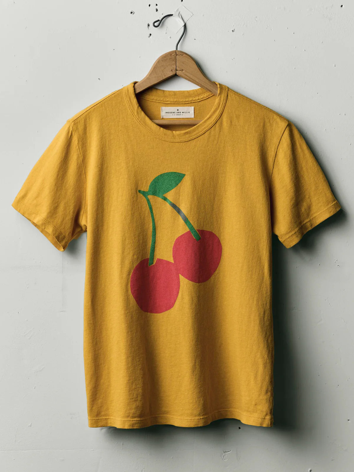 The Cherry Tee in Gold by Imogene + Willie on a wooden hanger, showing the full front with a large screen-printed cherry graphic — two red cherries with green stems and leaf — on a gold cotton tee