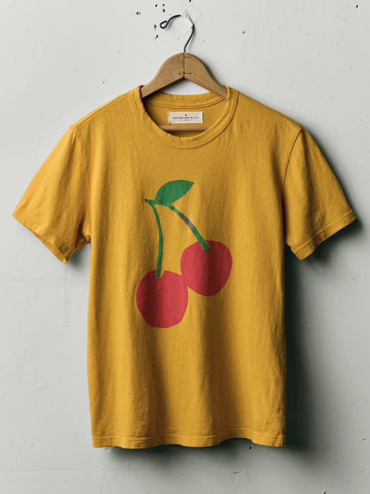The Cherry Tee in Gold by Imogene + Willie on a wooden hanger, showing the full front with a large screen-printed cherry graphic — two red cherries with green stems and leaf — on a gold cotton tee