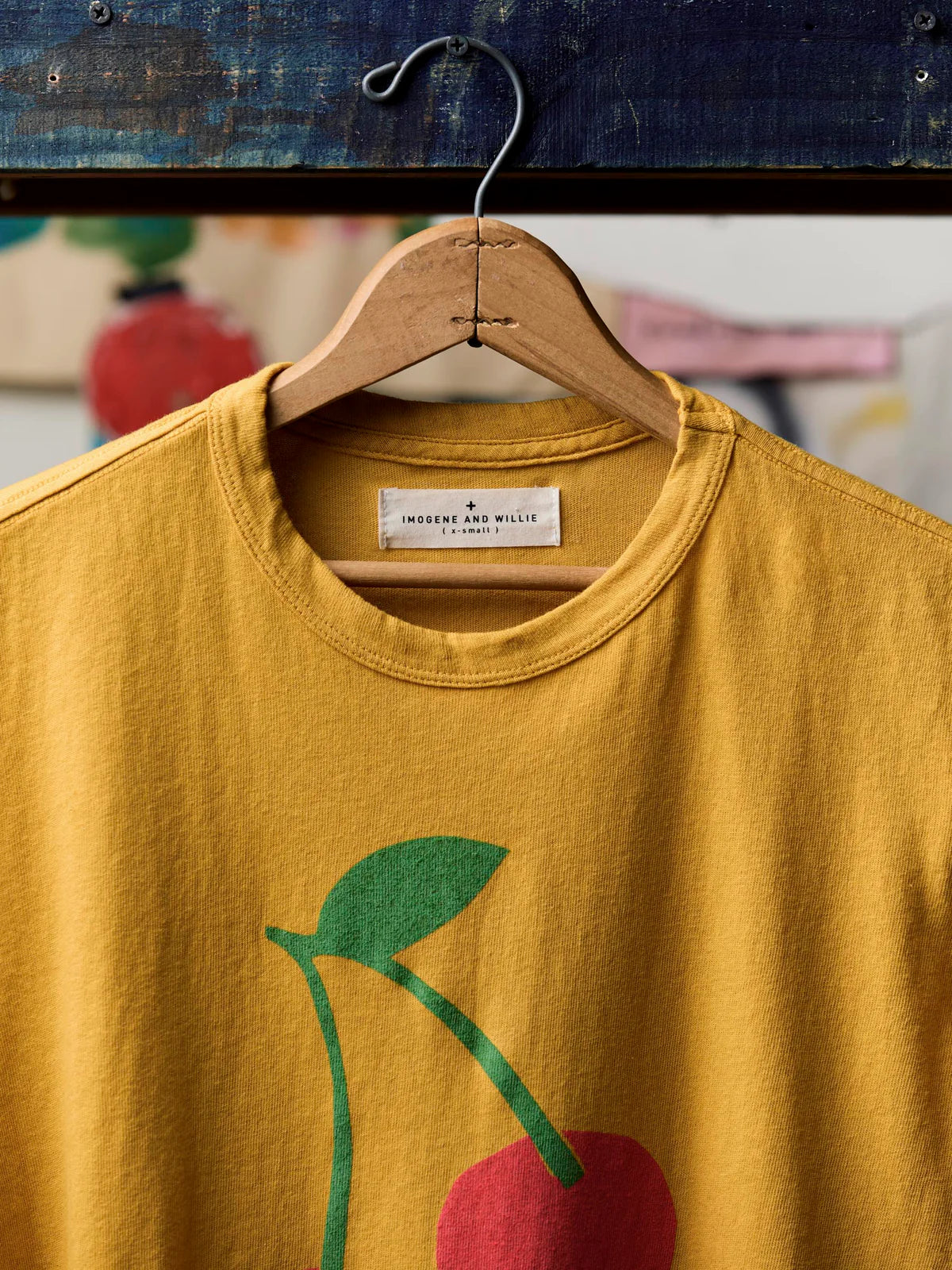 Close-up of the neckline of The Cherry Tee in Gold showing the Imogene and Willie label, ribbed crew neck, and the top of the cherry screen print with green stem and leaf on gold cotton