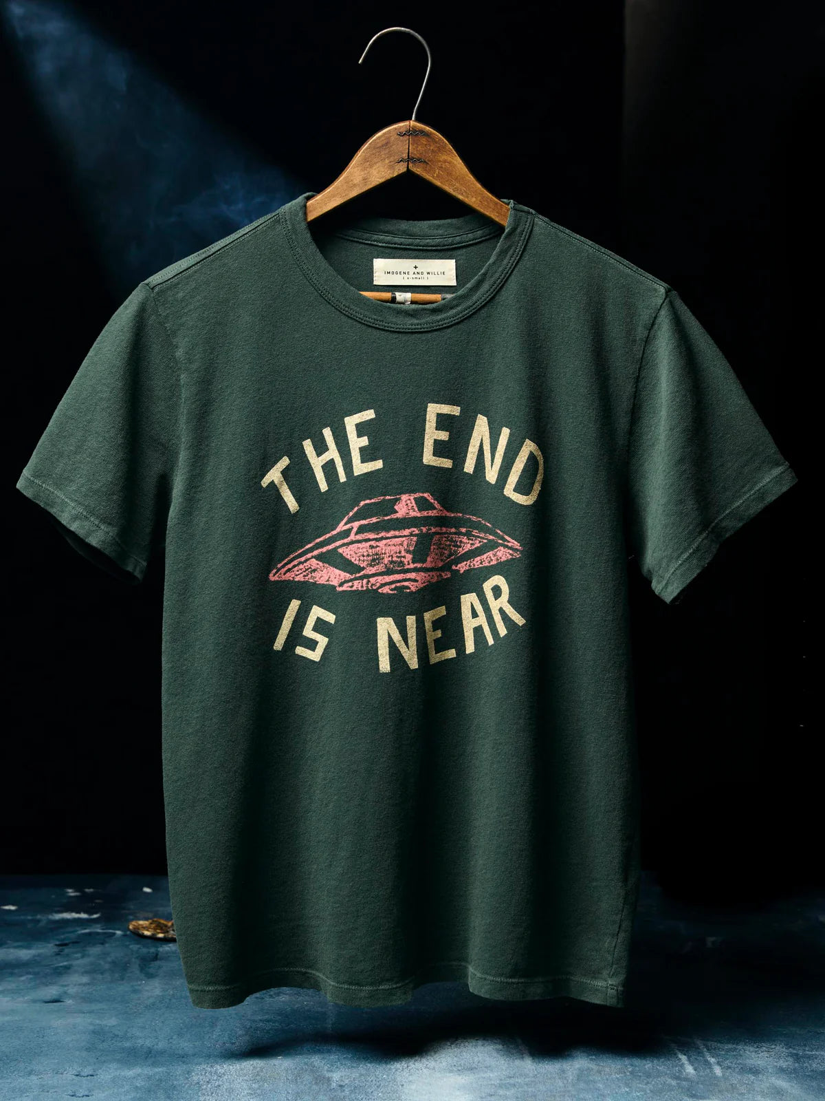 Dark spruce green short-sleeve tee on a wooden hanger, screen-printed with "The End Is Near" text and a vintage UFO graphic in cream and pink.
