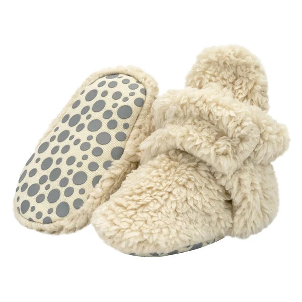 Oat faux-fur baby booties, with one tilted to show the dotted gripper sole and the other upright with its snap strap.
