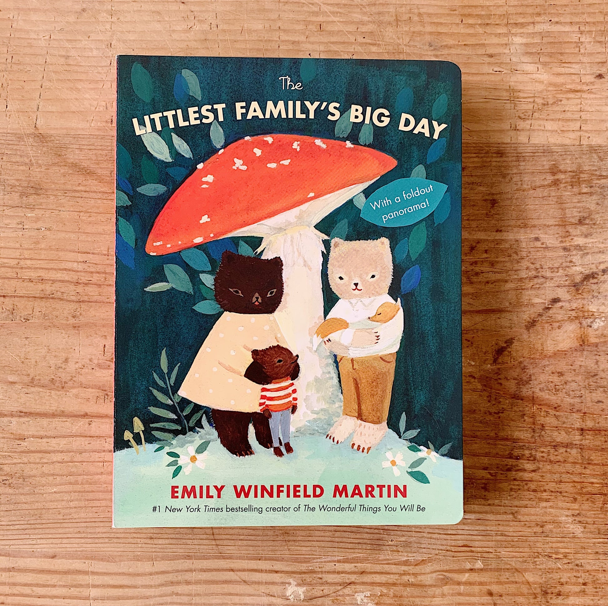 Cover of The Littlest Family's Big Day board book by Emily Winfield Martin, showing two illustrated bears and a small fox child sheltering under a large red mushroom, with teal foliage in the background and a badge reading "With a foldout panorama"
