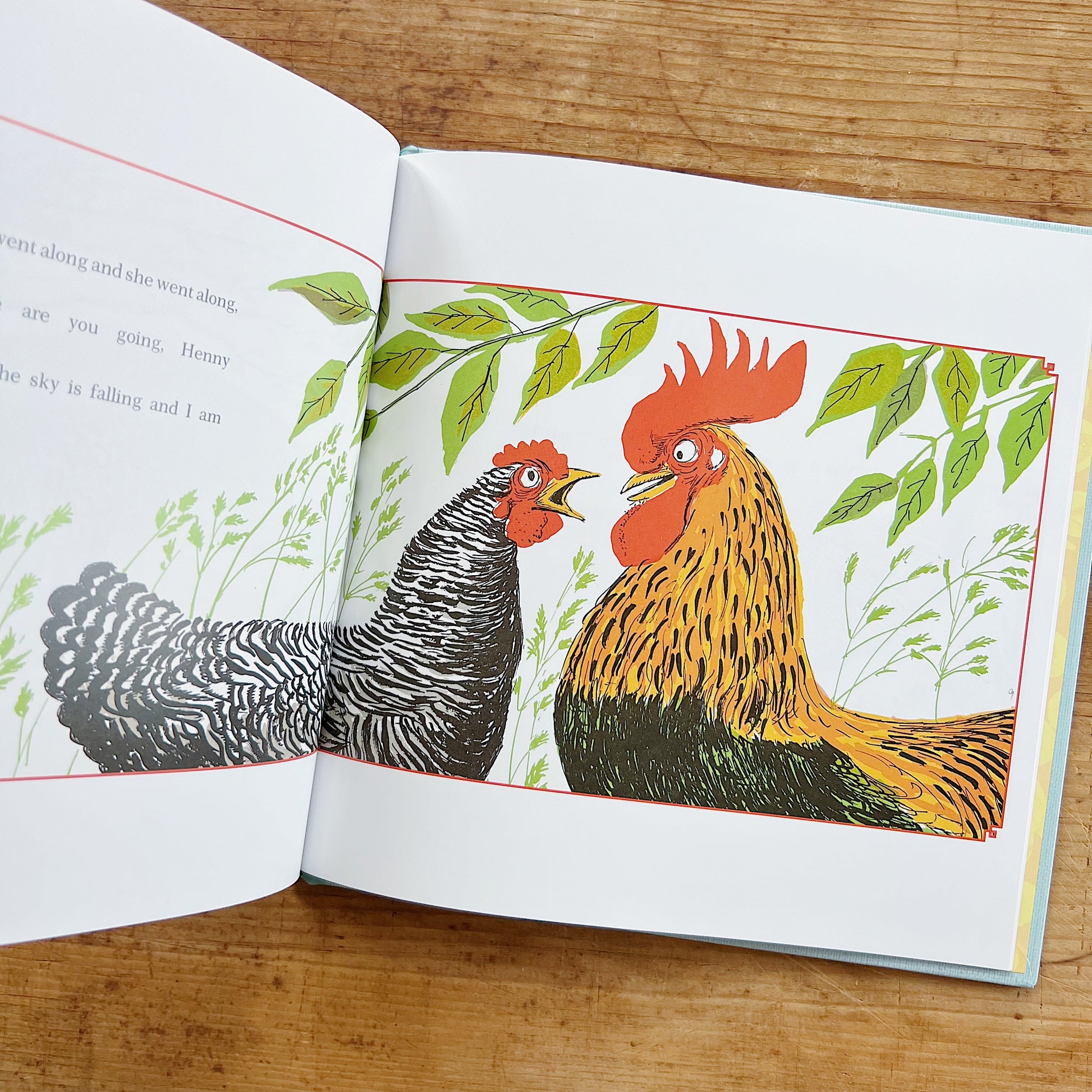 Open interior spread of Henny Penny showing story text on the left page with partial lines reading "went along and she went along," "are you going, Henny," and "he sky is falling and I am"; on the right, a full-page color illustration by Paul Galdone of Henny Penny, a barred black-and-white hen, facing Cocky Locky, a large golden-orange rooster with a vivid red comb, both standing amid tall green grasses and leafy branches, displayed on a warm wood surface.
