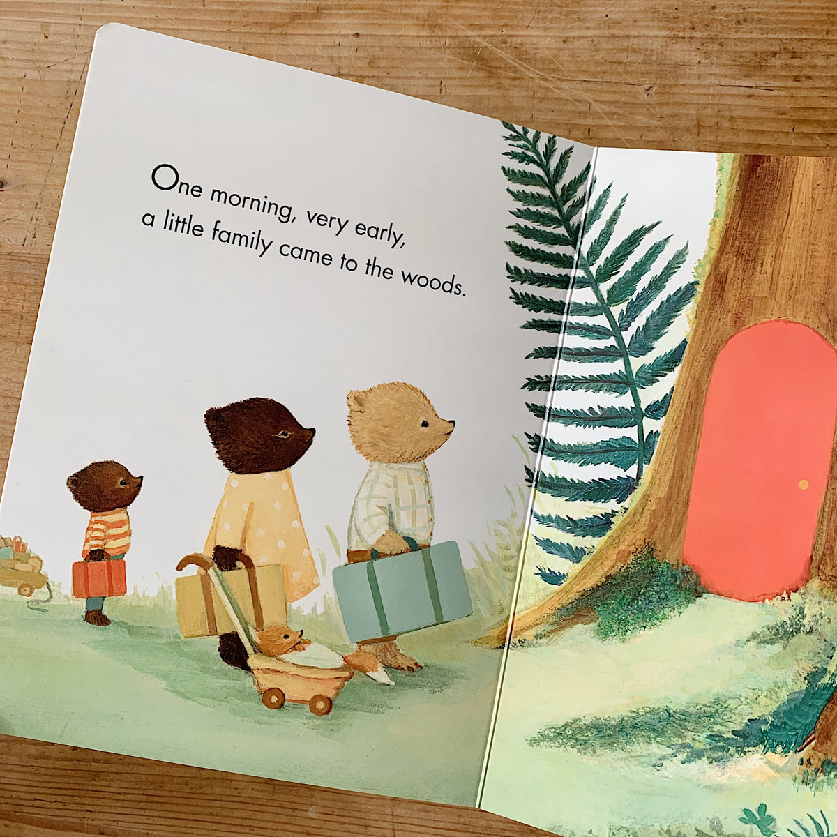 Interior spread of The Littlest Family's Big Day showing the bear family arriving at the woods with suitcases and a pram, beside text reading "One morning, very early, a little family came to the woods"
