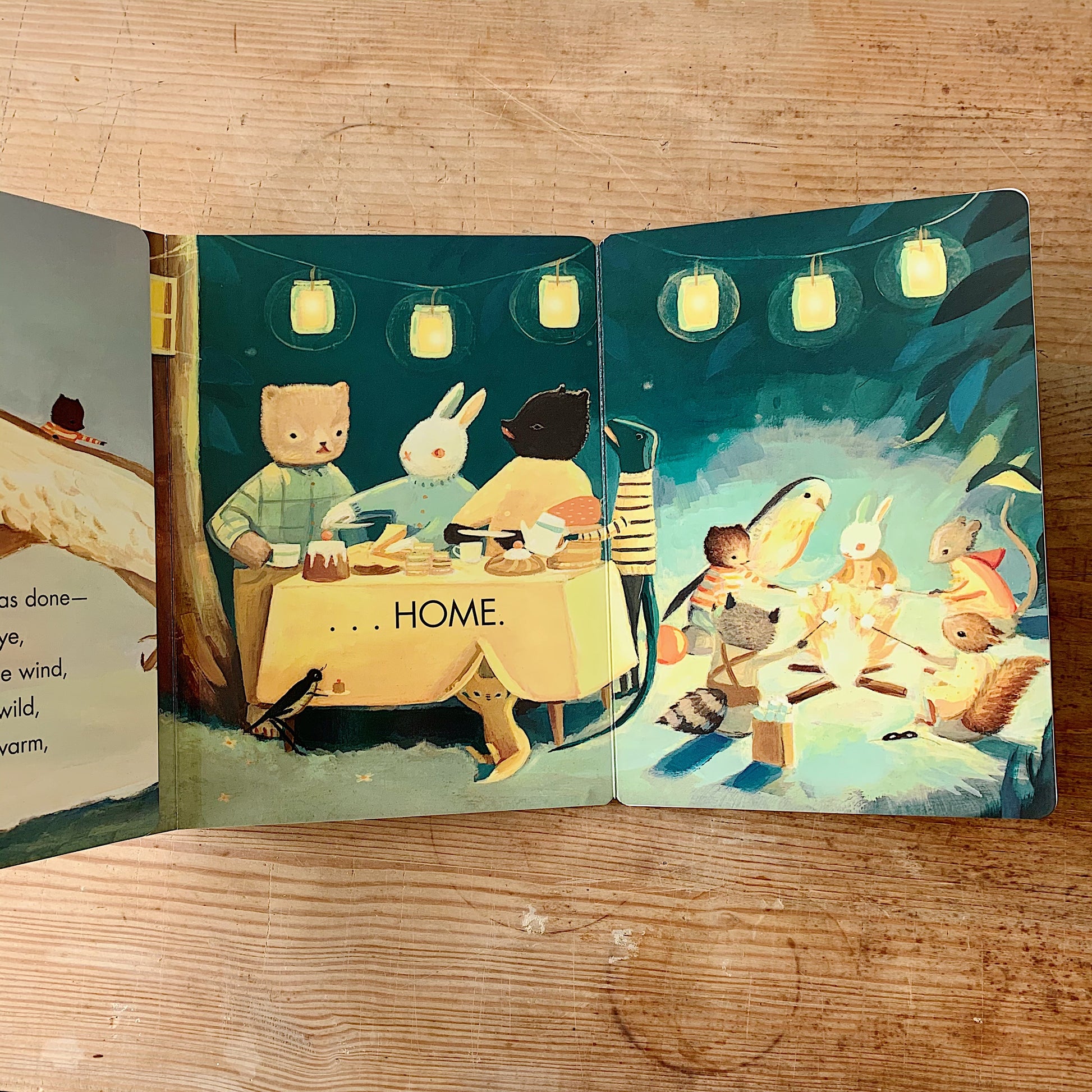 Interior spread of The Littlest Family's Big Day showing woodland animal characters gathered around a dinner table under glowing jar lanterns with the word HOME, and a campfire scene with animals toasting marshmallows on the right page
