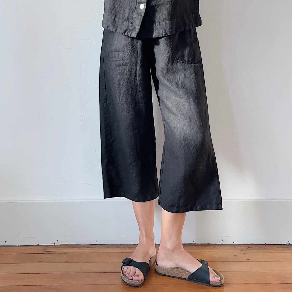 Model wearing Cut Loose Hanky Linen Easy Crop Flood in Black, front view showing black 100% linen wide-leg crop silhouette, front patch pockets, straight hem at mid-calf flood length, and natural linen texture, styled with matching black linen jacket and black Birkenstock sandals on wood floor against white wall
sandals