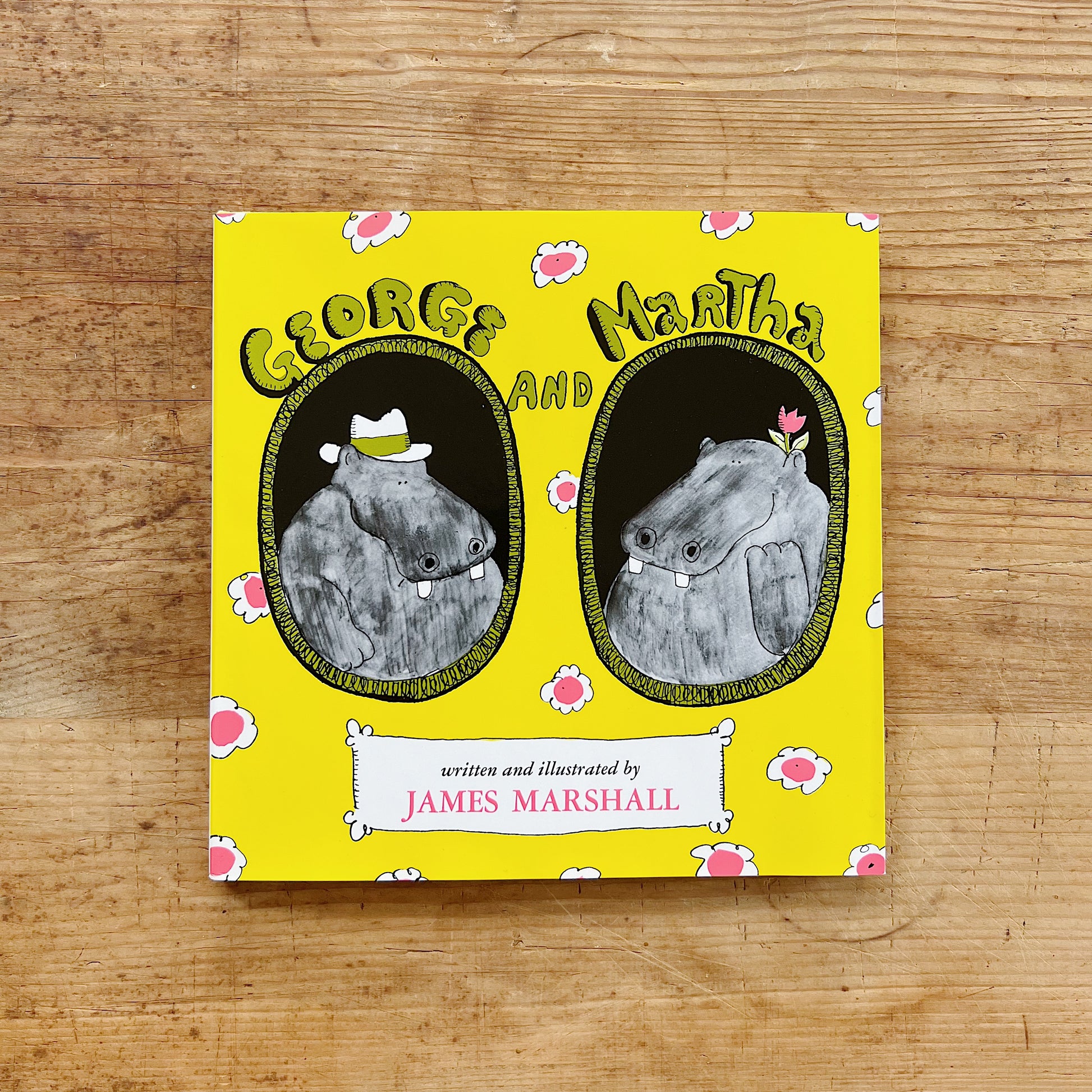 Front cover of George and Martha by James Marshall, featuring a bright yellow background scattered with pink daisy-like flowers, two oval portrait medallions side by side — on the left, George the hippo wearing a white fedora, and on the right, Martha the hippo with a small pink flower on her head — with the title in bubbly green lettering at the top and "written and illustrated by James Marshall" in a white banner at the bottom, displayed flat on a warm wood surface.
