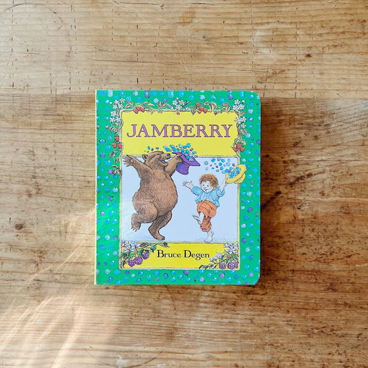 Front cover of Jamberry board book by Bruce Degen, featuring a bright green background scattered with iridescent blue and purple berry dots and a floral vine border of strawberries and white blossoms. A central framed panel on a light blue background shows a large brown bear rearing up on its hind legs and tossing a purple hat full of blueberries into the air, while a small red-haired boy in a blue shirt and orange trousers reaches up with a yellow hat to catch them. The title is printed in bold purple seri