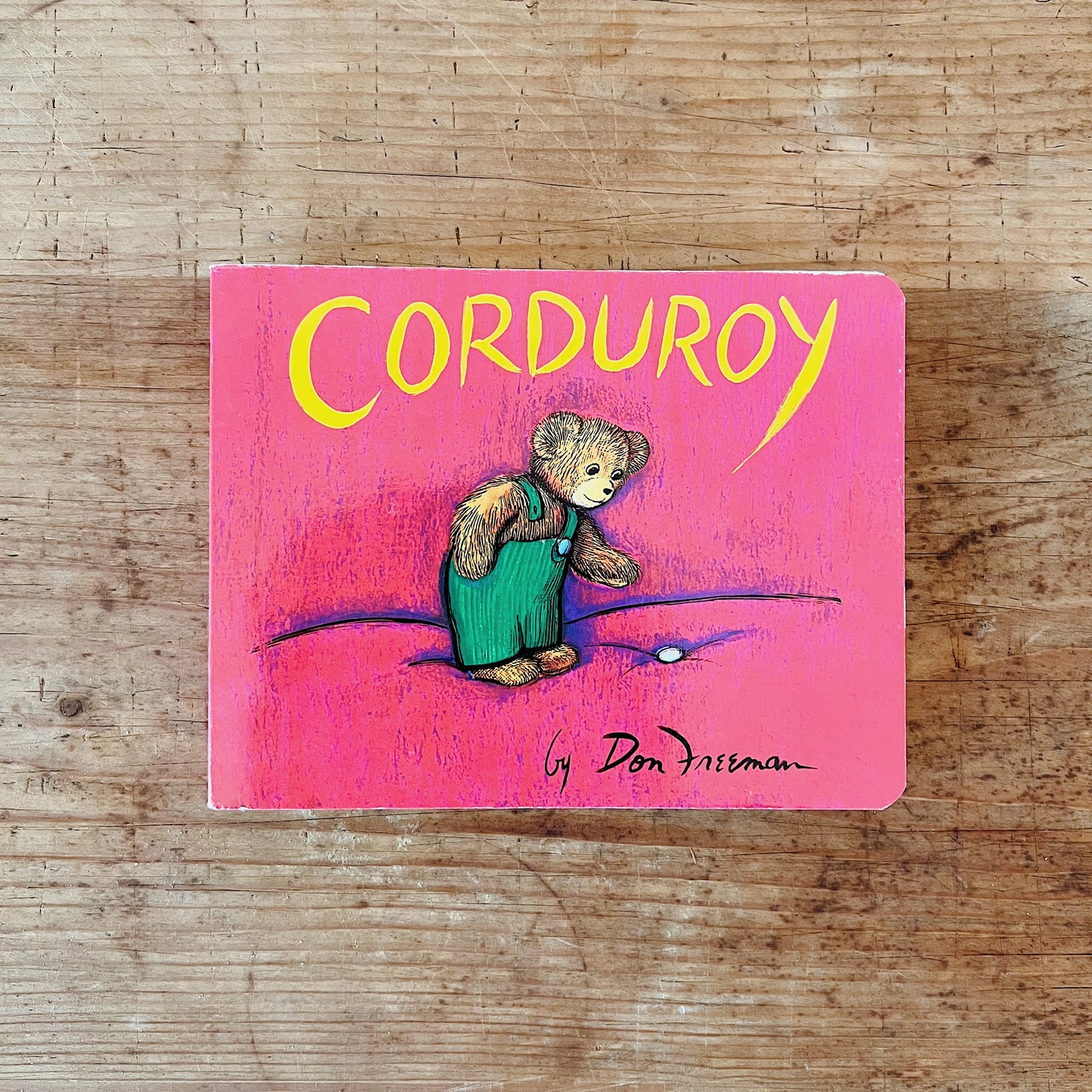 Front cover of Corduroy board book by Don Freeman, featuring a vivid hot pink background with the title in large rounded yellow lettering at the top and the author credit "by Don Freeman" in black script at the bottom. The central illustration shows Corduroy — a small brown teddy bear in green corduroy overalls — standing alone and looking downward toward a single white button lying on the ground in front of him, his expression wistful, displayed flat on a warm wood surface.
