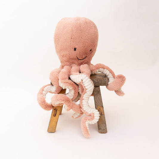 Jellycat Large Odell Octopus plush toy in blush pink, upright seated on a wooden stool showing a smiling face and eight dangling tentacles with cream sucker details on white background