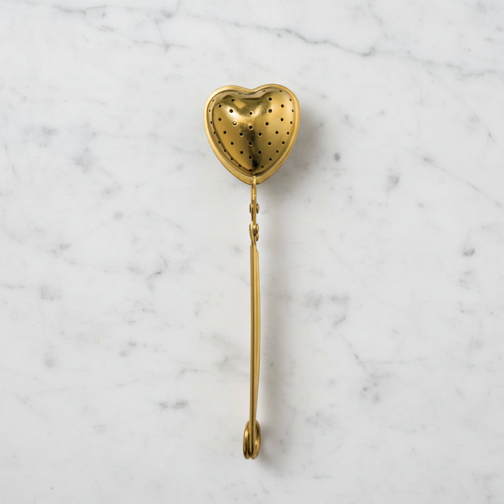 Gold-finish stainless steel heart-shaped tea strainer with squeeze handle, laid flat on a white marble surface