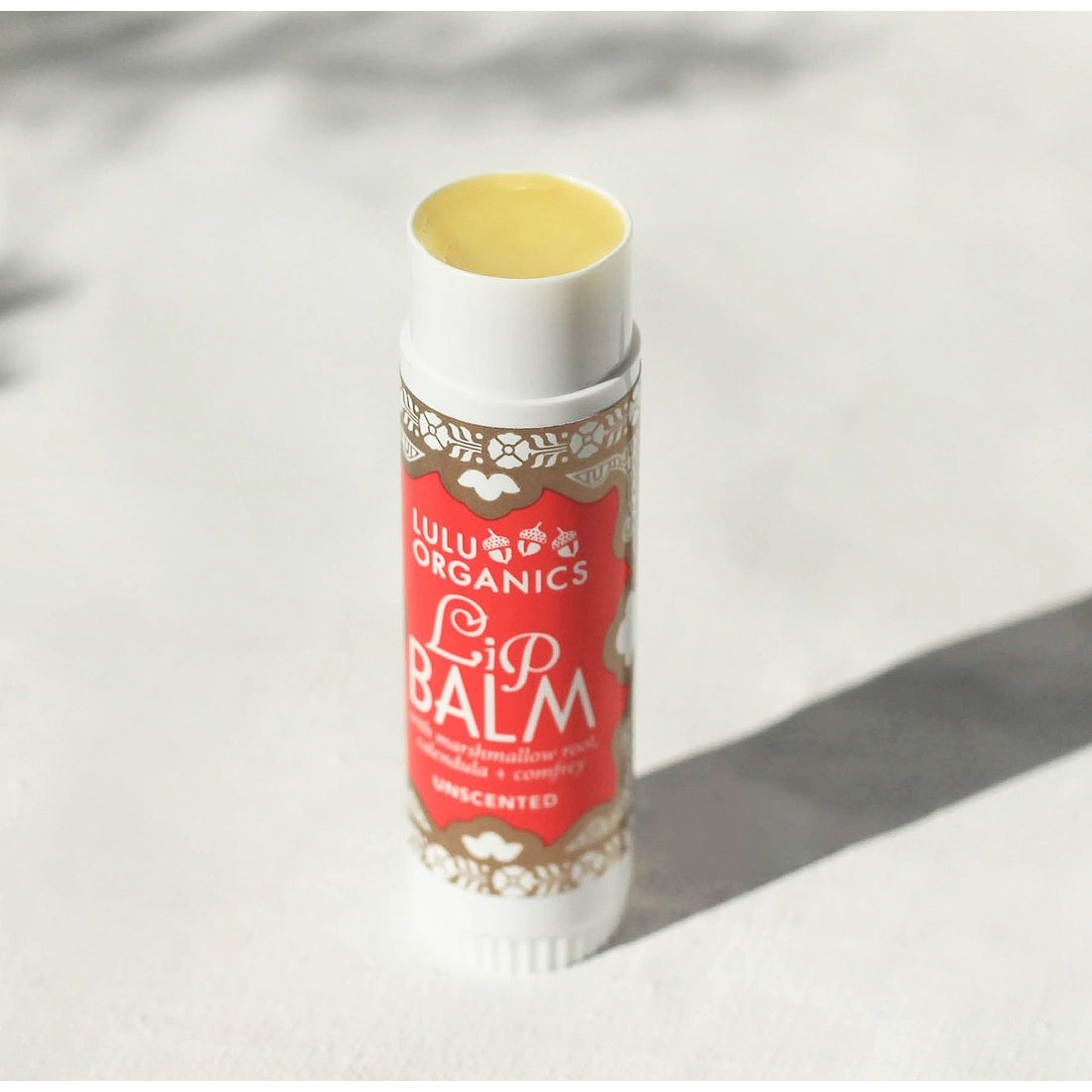 Lulu Organics Unscented Big Lip Balm open tube standing upright showing yellow balm, with red and white illustrated label reading Lip Balm with marshmallow root, calendula and comfrey, Unscented, on a white stone surface in sunlight

